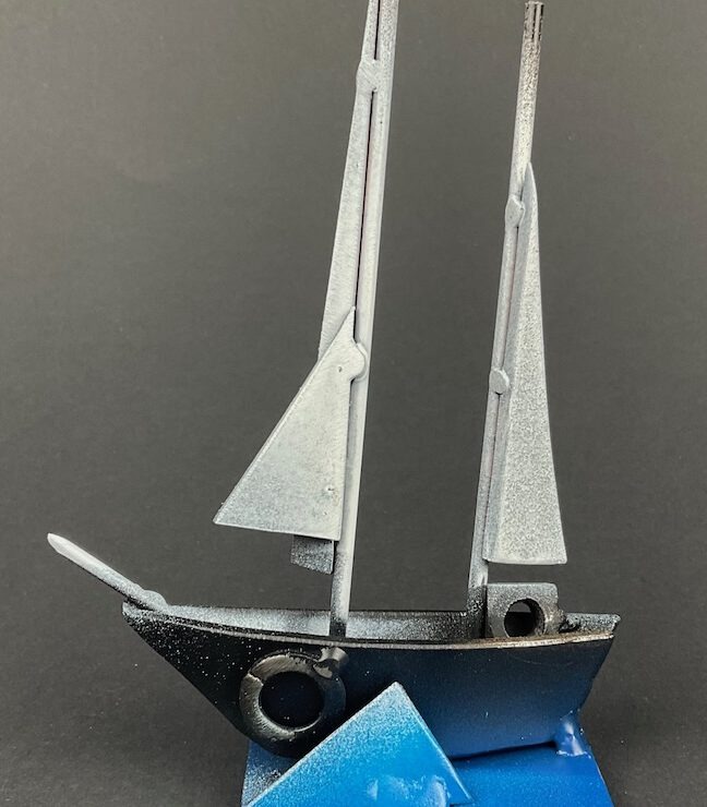 Eine moderne Metallskulptur eines Segelbootes mit zwei Masten und Segeln, die mit weißer und silberner Farbe besprüht sind. Der Rumpf ist dunkel und steht auf einem geometrischen blauen Metallockel, der Wellen symbolisiert. Der Hintergrund ist dunkelgrau.