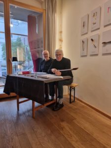 Lichtdurchfluteter Raum mit Holzboden. Zwei Männer sitzen an einem Holztisch mit schwarzer Decke. Der Mann links trägt eine Brille und einen dunklen Pullover. Der Mann rechts, ebenfalls mit Brille, trägt einen schwarzen Pullover, eine dunkle Hose mit gemusterten Socken und dunkle Schuhe. Beide schauen lächelnd in die Kamera. Der Mann rechts hält einen Gegenstand in der Hand und schaut lächelnd in die Kamera. Hinter ihnen befindet sich eine Wand mit gerahmten Kunstwerken. Links ist ein großes Fenster mit Blick nach draußen.